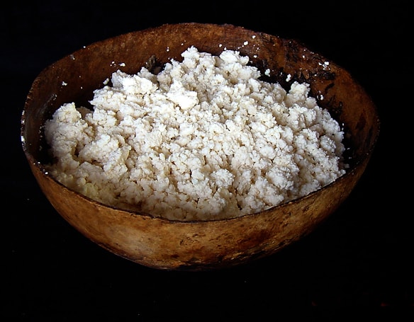 A rustic wooden bowl filled with white, crumbly cheese or a similar dairy product, sitting on a dark background. The texture of the cheese is visible, with small clumps and a slightly rough surface.