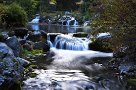 A serene natural landscape featuring a cascading waterfall flowing over rocks into a tranquil pond. Lush greenery surrounds the area, with trees and bushes adding to the tranquil environment. The water appears smooth and reflective, capturing the peaceful essence of the scene.