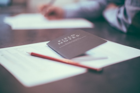 A notebook with 'Field Notes' written on the cover is placed on a table alongside a few sheets of paper and a pencil. The background is slightly blurred, showing a person possibly writing or reviewing documents.