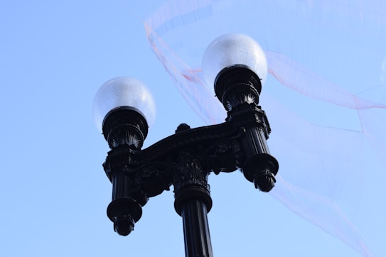 A vintage-style street lamp post rises against a clear blue sky. The lamp features two spherical, frosted glass lights with an ornate, black metal design. A thin, translucent fabric flows behind it, adding a dynamic element.