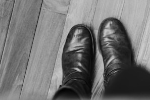 A minimalist black and white photo of men's boots placed on a dark floor.