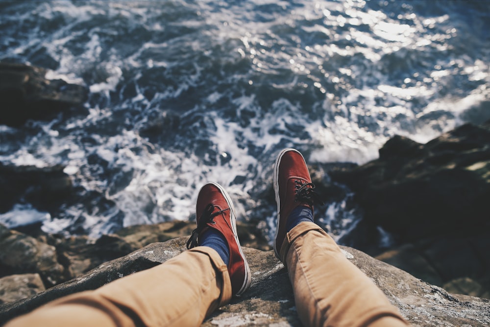 man in red sneakers sitting in cliff near body of water