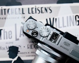 The photograph features a Fujifilm camera placed over what appears to be a vintage movie poster. The camera's dials and lens are prominently in view. The poster underneath displays several text elements such as 'Mitchell Leisen's Production' and contains silhouettes of a person wearing a hat.