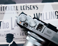 The photograph features a Fujifilm camera placed over what appears to be a vintage movie poster. The camera's dials and lens are prominently in view. The poster underneath displays several text elements such as 'Mitchell Leisen's Production' and contains silhouettes of a person wearing a hat.