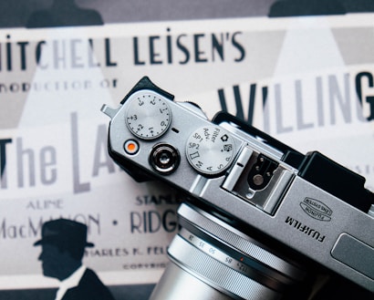 The photograph features a Fujifilm camera placed over what appears to be a vintage movie poster. The camera's dials and lens are prominently in view. The poster underneath displays several text elements such as 'Mitchell Leisen's Production' and contains silhouettes of a person wearing a hat.