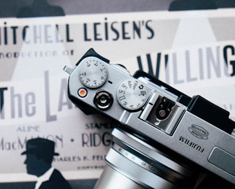 The photograph features a Fujifilm camera placed over what appears to be a vintage movie poster. The camera's dials and lens are prominently in view. The poster underneath displays several text elements such as 'Mitchell Leisen's Production' and contains silhouettes of a person wearing a hat.