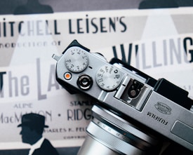 The photograph features a Fujifilm camera placed over what appears to be a vintage movie poster. The camera's dials and lens are prominently in view. The poster underneath displays several text elements such as 'Mitchell Leisen's Production' and contains silhouettes of a person wearing a hat.