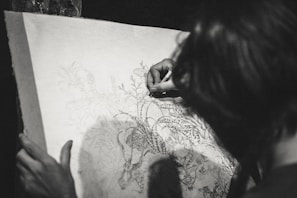 A person is engaged in a detailed drawing, focusing intently on the artwork. The drawing features intricate designs of flora and fauna, suggesting a theme of nature. The image is in black and white, enhancing the focus on the pencil work and the concentration of the artist.