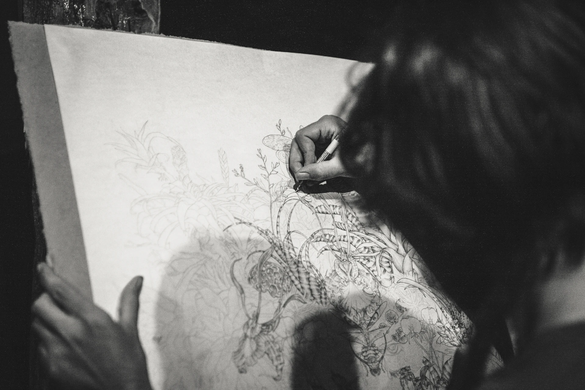 Black-and-white photograph of a person sketching an intricate botanical tattoo design on paper.
