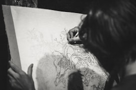 A person is engaged in a detailed drawing, focusing intently on the artwork. The drawing features intricate designs of flora and fauna, suggesting a theme of nature. The image is in black and white, enhancing the focus on the pencil work and the concentration of the artist.