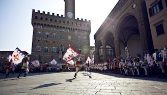 A group of people in historical costumes engage in a lively parade in a large piazza. They are holding colorful flags featuring a red emblem. The background shows a historic stone building with a clock tower, likely a famous architectural landmark. A large crowd lines the sides, watching the event.