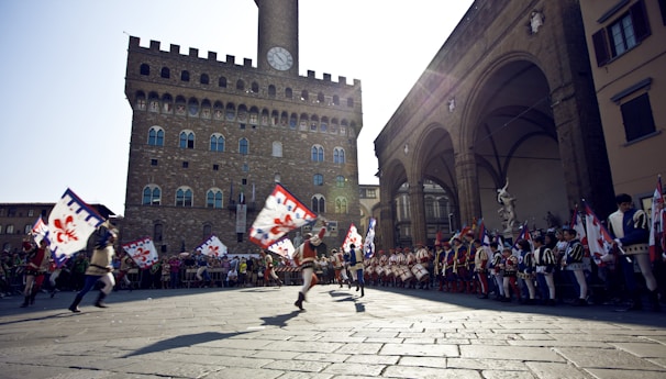 A group of people in historical costumes engage in a lively parade in a large piazza. They are holding colorful flags featuring a red emblem. The background shows a historic stone building with a clock tower, likely a famous architectural landmark. A large crowd lines the sides, watching the event.