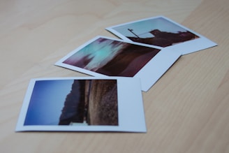 Three polaroid photographs are scattered on a light wooden surface. Each photograph depicts different landscapes or scenes with a nostalgic and vintage appearance due to their color tone and composition.