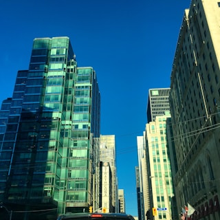 A cityscape featuring tall modern buildings with reflective glass windows under a clear blue sky. The streets below are partially visible with vehicles and a street sign indicating traffic directions.