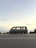 A van is parked on a road beside a scenic seaside landscape, with a calm ocean and clear sky in the background. The vehicle appears to be stationary, situated on an empty road bordered by greenery.