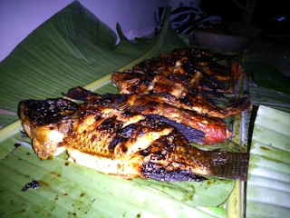 Several grilled fish are arranged on a banana leaf, showcasing charred and crispy skin with a golden-brown color. The setting appears to be outdoors or in a rustic kitchen with minimal lighting.