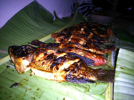 Several grilled fish are arranged on a banana leaf, showcasing charred and crispy skin with a golden-brown color. The setting appears to be outdoors or in a rustic kitchen with minimal lighting.