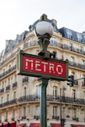 A classic Parisian metro sign with art deco design stands in front of a traditional Haussmann-style building. The sign is red with white letters, and it is topped with an ornate street lamp. The building in the background features multiple windows and wrought-iron balconies.