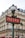 A classic Parisian metro sign with art deco design stands in front of a traditional Haussmann-style building. The sign is red with white letters, and it is topped with an ornate street lamp. The building in the background features multiple windows and wrought-iron balconies.