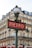 A classic Parisian metro sign with art deco design stands in front of a traditional Haussmann-style building. The sign is red with white letters, and it is topped with an ornate street lamp. The building in the background features multiple windows and wrought-iron balconies.