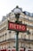 A classic Parisian metro sign with art deco design stands in front of a traditional Haussmann-style building. The sign is red with white letters, and it is topped with an ornate street lamp. The building in the background features multiple windows and wrought-iron balconies.
