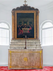 A religious setting features a prominent altar with a decorative gold emblem on the front. Above the altar is a large framed painting depicting a historic scene with several figures gathered around a table, reminiscent of a famous religious artwork. The setting includes ornate architectural details and a central cross.