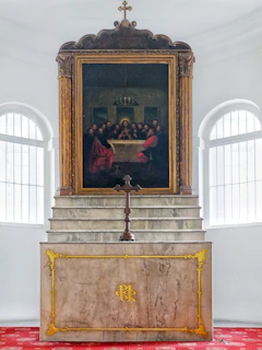 A religious setting features a prominent altar with a decorative gold emblem on the front. Above the altar is a large framed painting depicting a historic scene with several figures gathered around a table, reminiscent of a famous religious artwork. The setting includes ornate architectural details and a central cross.