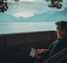 man sitting while holding a book watching on body of water