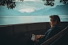 man sitting while holding a book watching on body of water