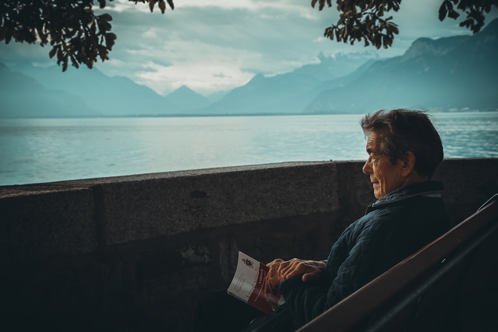hombre sentado mientras sostiene un libro mirando en el cuerpo de agua