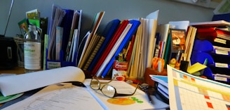 A cluttered office desk with various documents, folders, and stationery items scattered around. There are multiple colored binders and a can of Campbell's soup prominently placed. A pair of reading glasses rests on top of some papers, and there are notes and a calendar visible. A bottle and several pens are also present.