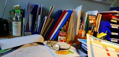 A cluttered office desk with various documents, folders, and stationery items scattered around. There are multiple colored binders and a can of Campbell's soup prominently placed. A pair of reading glasses rests on top of some papers, and there are notes and a calendar visible. A bottle and several pens are also present.