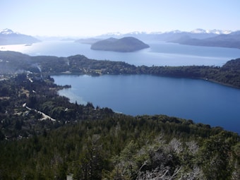 A serene landscape featuring an expansive lake surrounded by lush, green forests and distant mountains with snow-capped peaks. The water's surface reflects the clear blue sky, and scattered islands are visible in the distance beneath the soft, natural lighting.