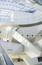 A modern, white and sleek architectural interior features multiple levels interconnected by glass railings. The space is illuminated by natural light streaming through a large, geometric glass ceiling. Clean lines and minimalistic design elements dominate the scene.