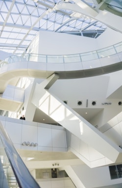 A modern, white and sleek architectural interior features multiple levels interconnected by glass railings. The space is illuminated by natural light streaming through a large, geometric glass ceiling. Clean lines and minimalistic design elements dominate the scene.