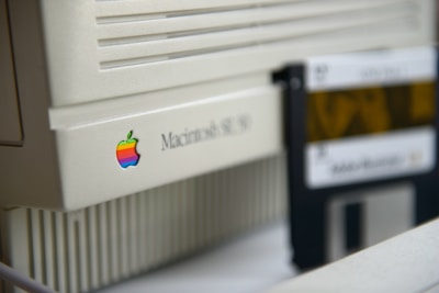 A close-up view of a vintage Macintosh SE/30 computer, displaying the iconic colorful Apple logo. To the right, there is a floppy disk labeled as Adobe Illustrator 3.0, suggesting a retro technology theme.