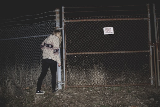 A person stands near a chain-link fence with a sign that reads 'Private Property No Trespassing.' The scene is set at night, with the area dimly lit, creating a mysterious atmosphere. The person is wearing a patterned sweater and black pants, and the ground is covered in dry grass.