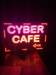 Brightly lit sign displaying the words 'CYBER CAFE' with an arrow pointing to the right, using colorful LED lights against a dark background.