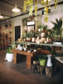 A cozy interior of a plant and pottery shop with wooden tables displaying an array of ceramic pots and lush green plants. The ceiling is adorned with hanging golden decorations, enhancing the warm and stylish decor. Shelves housing additional plants and decorative items line the walls, contributing to the inviting atmosphere.