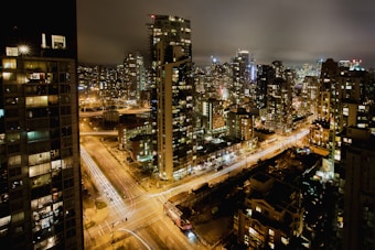 A vibrant cityscape at night, filled with tall buildings illuminated by an array of lights. Streets glow with the motion blur of traffic, and the skyline is shrouded by a foggy night sky.