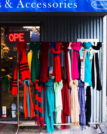 A display of colorful scarves hangs in front of a shop. The scarves feature a variety of patterns and textures in vibrant colors like red, pink, blue, green, and beige. Behind them, the shop's glass entrance shows a neon 'OPEN' sign, and the interior of the shop is slightly visible. The setting conveys a welcoming and lively vibe.