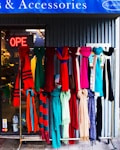 A display of colorful scarves hangs in front of a shop. The scarves feature a variety of patterns and textures in vibrant colors like red, pink, blue, green, and beige. Behind them, the shop's glass entrance shows a neon 'OPEN' sign, and the interior of the shop is slightly visible. The setting conveys a welcoming and lively vibe.