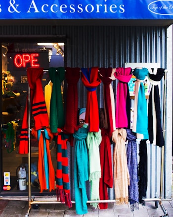A display of colorful scarves hangs in front of a shop. The scarves feature a variety of patterns and textures in vibrant colors like red, pink, blue, green, and beige. Behind them, the shop's glass entrance shows a neon 'OPEN' sign, and the interior of the shop is slightly visible. The setting conveys a welcoming and lively vibe.