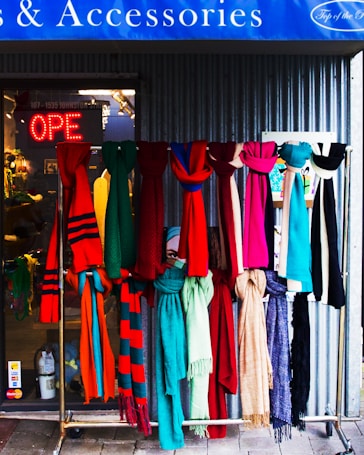 A display of colorful scarves hangs in front of a shop. The scarves feature a variety of patterns and textures in vibrant colors like red, pink, blue, green, and beige. Behind them, the shop's glass entrance shows a neon 'OPEN' sign, and the interior of the shop is slightly visible. The setting conveys a welcoming and lively vibe.