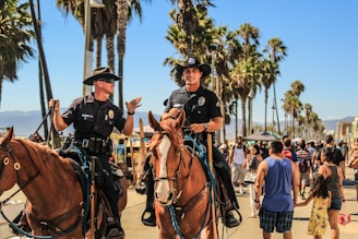Two police officers on horseback patrol a busy boardwalk lined with palm trees under a clear, sunny sky. They are wearing uniforms and wide-brimmed hats, and are engaged in conversation. People walk by, including a man in a blue tank top holding a child's hand.