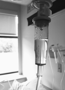 A close-up view of an intravenous drip chamber filled with liquid, in a clinical setting with hospital equipment and a window visible in the background. The image is in black and white, emphasizing the medical and sterile environment.