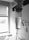 A close-up view of an intravenous drip chamber filled with liquid, in a clinical setting with hospital equipment and a window visible in the background. The image is in black and white, emphasizing the medical and sterile environment.