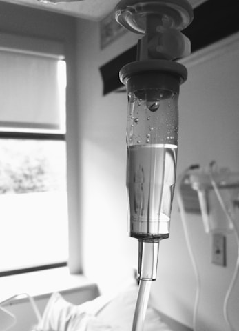 A close-up view of an intravenous drip chamber filled with liquid, in a clinical setting with hospital equipment and a window visible in the background. The image is in black and white, emphasizing the medical and sterile environment.