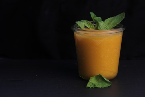 A glass jar filled with a vibrant orange smoothie, topped with fresh mint leaves. The jar is placed on a dark surface with a few mint leaves scattered nearby. The background is black, highlighting the brightness of the smoothie.