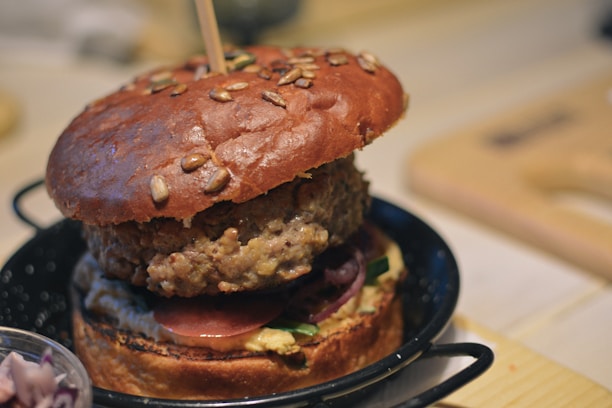 A close-up of a gourmet hamburger featuring a large, juicy beef patty sandwiched between two halves of a seed-topped bun. The burger includes fresh ingredients such as red onions, tomato slices, and what appears to be a sauce or spread on the toasted bottom bun. The burger is served in a small black skillet with handles.